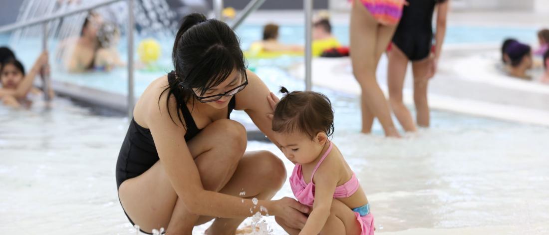 A mother crouches near her toddler at a shallow pool