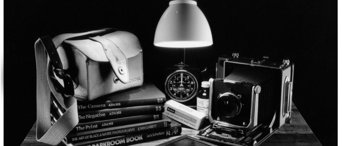 Black and white photograph showing objects including a lamp, wallet, and camera on a wooden table.
