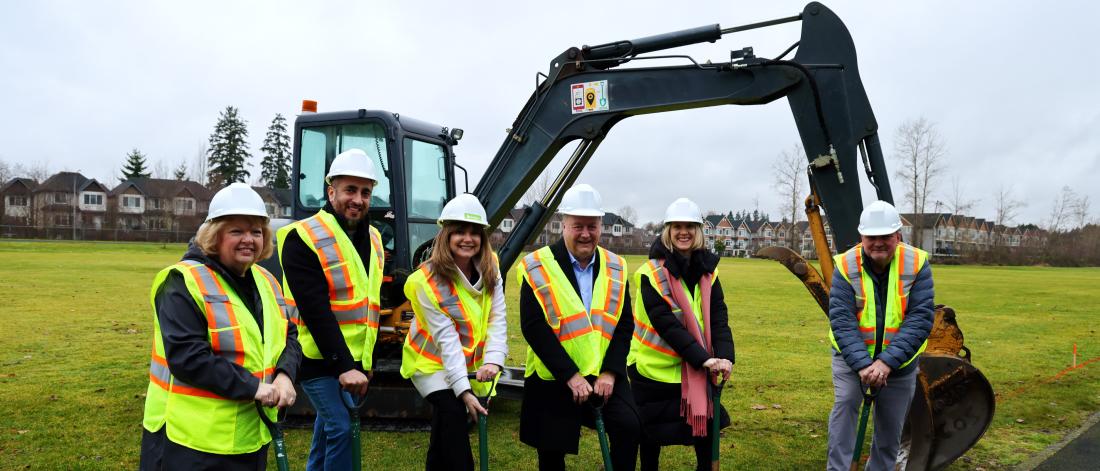 Mayor and Council use shovels to break ground beside field in front of excavator