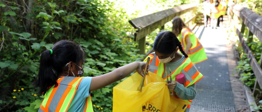 Two children help pick up litter along a forest path.