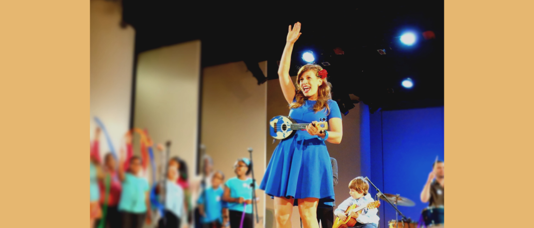 Mill wears a blue dress and holds a blue ukulele and waves at the audience