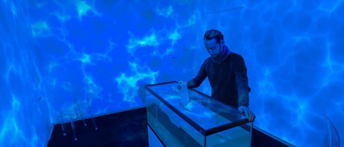 Person inserts handwritten letter into water tank in a water-inspired room at an art gallery.