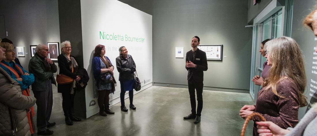 Rhys Edwards leads an exhibition tour in Surrey Art Gallery, surrounded by circle of people listening.