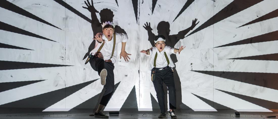 Two performers for show Doodle POP stand on stage with arms outstretched in the centre of a background projection of black and white lines.
