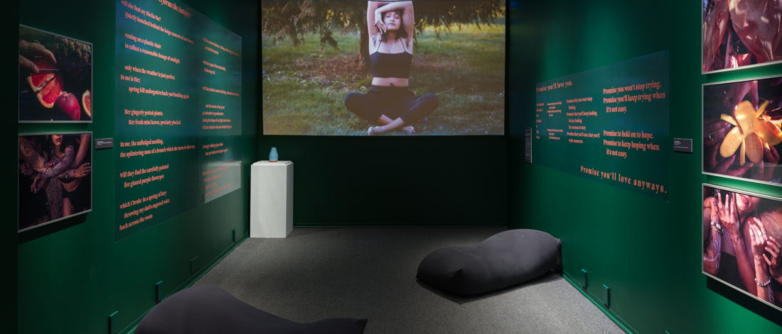 Two green walls with text and photographs frame a space with two yogibos. A film plays on the wall in between the two greens where a woman sitting cross-legged on grass in nature is shown. She has her elbows bent overhead, one hand cups her jaw, the other hand is behind her back.