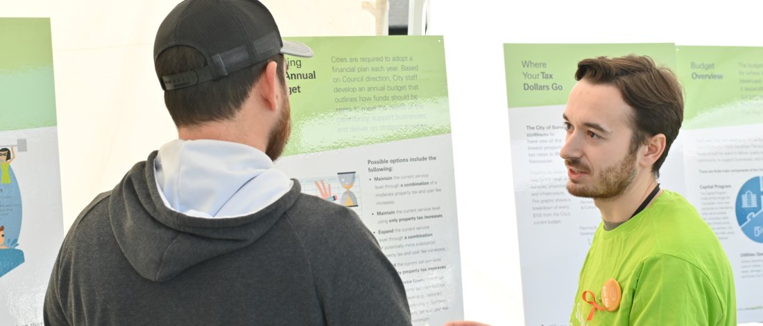Two men, one in a green shirt and the other in a grey hoodie, are discussing information on posters about municipal budgeting at a public event.