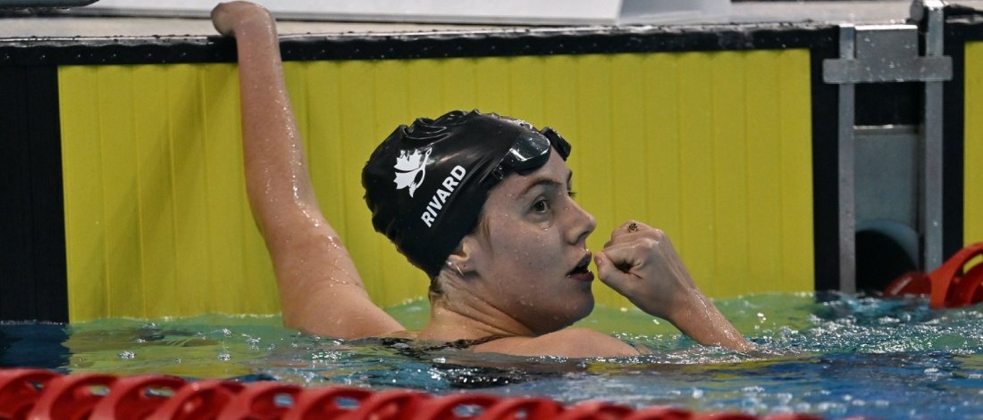  A swimmer with "RIVARD" on their cap looks aside at the pool's edge, goggles on their forehead.
