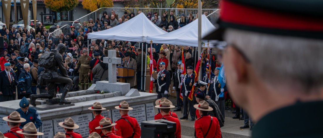 A Remembrance Day ceremony with uniformed individuals in red jackets and attendees around a soldier statue paying respect.