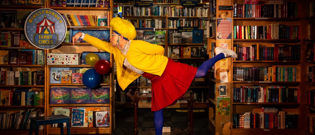 the librarian wearing a yellow cardigan, red skirt and blue tights is in the library in front of bookshelves with lots of books, is balancing on one leg with the other let outstretched and arm in the air