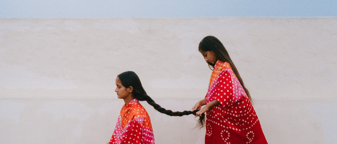Two children face the left side of the photograph. One child stands behind the seated one to braid her long hair. They both wear bright pink, orange, pink printed clothing, against a white backdrop.