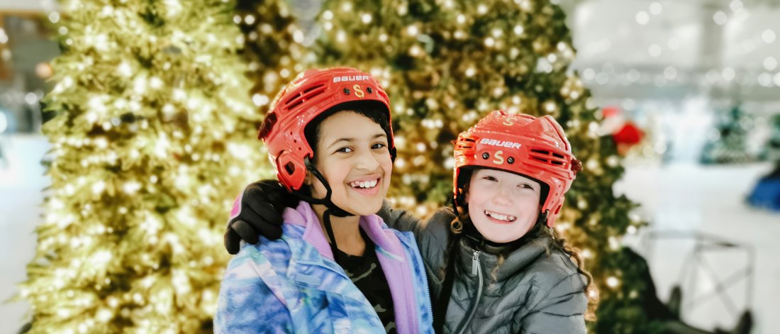 Two smiling kids in skating gear pose before a lit-up Christmas tree.