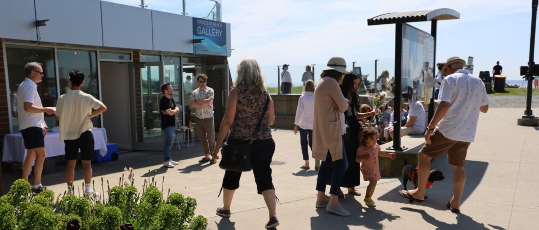 People socializing on a sunny day outside the Crescent Beach Gallery, with flowers in the foreground.