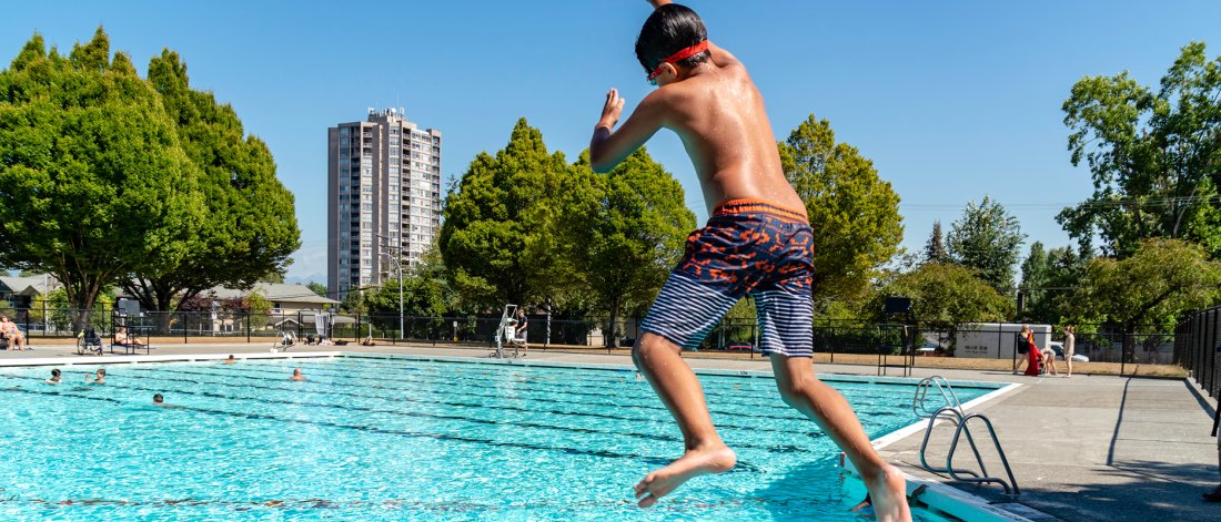 A boy jumping into an outdoor pool with trees and a building in the background.