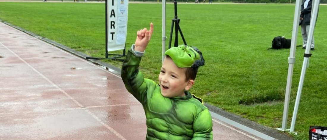  A joyful child in a green costume and helmet, raising a hand in excitement, seated in a racing wheelchair on a wet track..