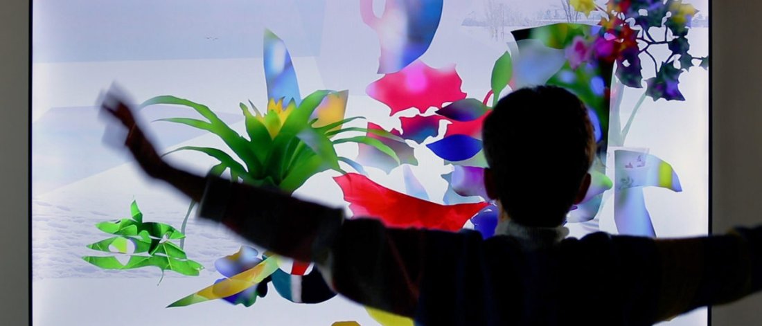 A person with their arms out-wide is in front of an interactive exhibit. Plant shapes of green, red, yellow, blue and purple colours can be seen against a pale blue screen that suggest snow and the outdoors.