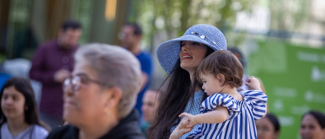 A smiling woman in a blue sun hat holding a young child at an outdoor event, with people in the background.