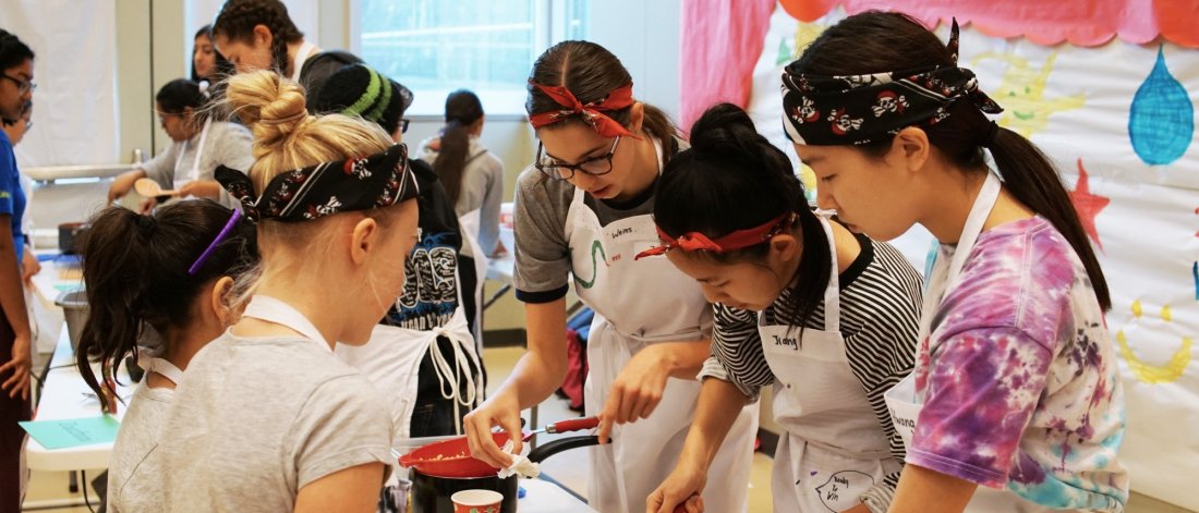 A cooking workshop with young people of various ages, wearing aprons and bandanas, actively engaged in preparing food.