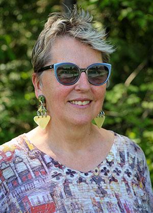 Photo of Jude Campbell looking at the camera, a woman with dark sunglasses, short white-blonde hair, wearing a multi-colored shirt and gold earrings.