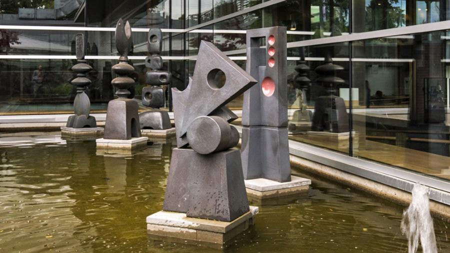 Five grey vertical sculptures made of clay in various geometric and organic stand on separate bases in a reflecting pool.
