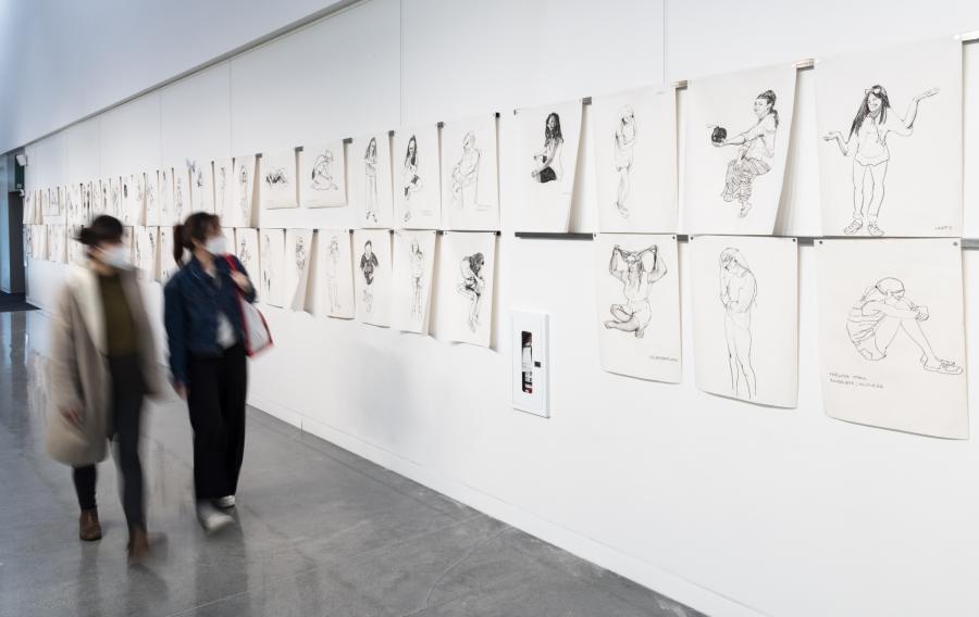 A long gallery wall with life-drawing images hung on it and two people walking towards the camera, looking at the drawings.
