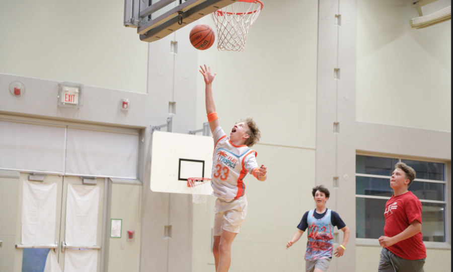 Boy in a white and orange jersey doing a layup with a basketball with other boys ready for the rebound