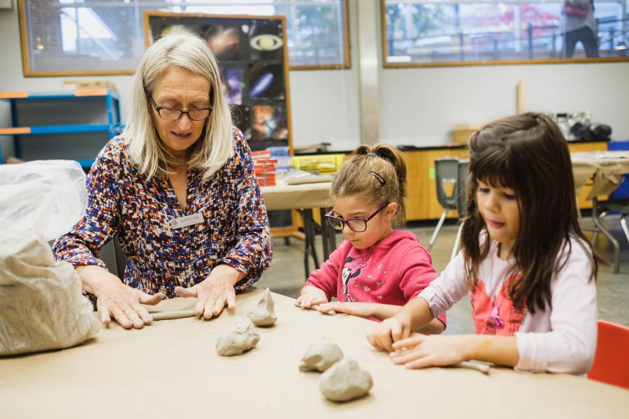 A teacher and two kids sit at a table in a classroom rolling out clay