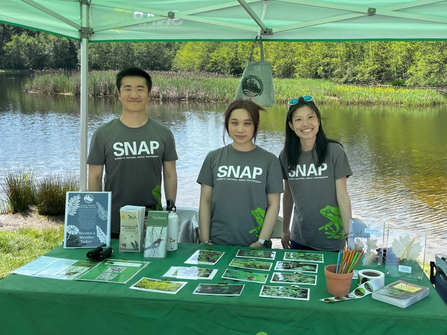 three people standing by a lake, behind a table with brochures and photos on it