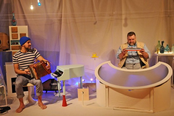 two performers on stage, one sits at a tiny piano holding an accordion, the other sits behind a crescent moon shaped table holding a small boat