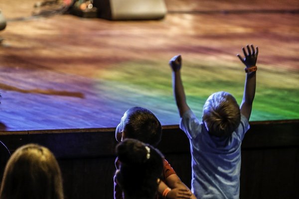 view is behind a young boy in the front row of the theatre who has his hands up in the air enjoying a performance on stage 