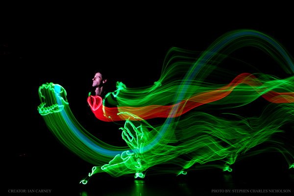 black background you see a performer's face amongst swirling lights of green, red and blue in the shape of a dinosaur 