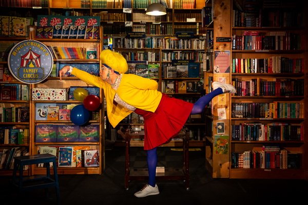 the libravian performer with a bright yellow wig, bright yellow sweater and red skirt in front of the library set standing on one foot with a leg and arm outstretched. 