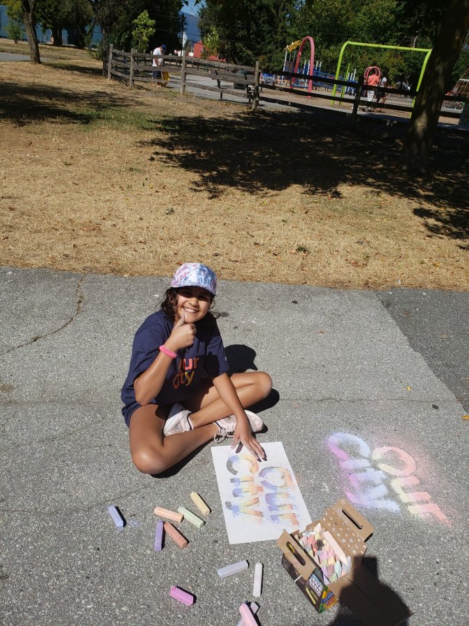A smiling young girl beside her chalk art on the pavement in a sunny park.