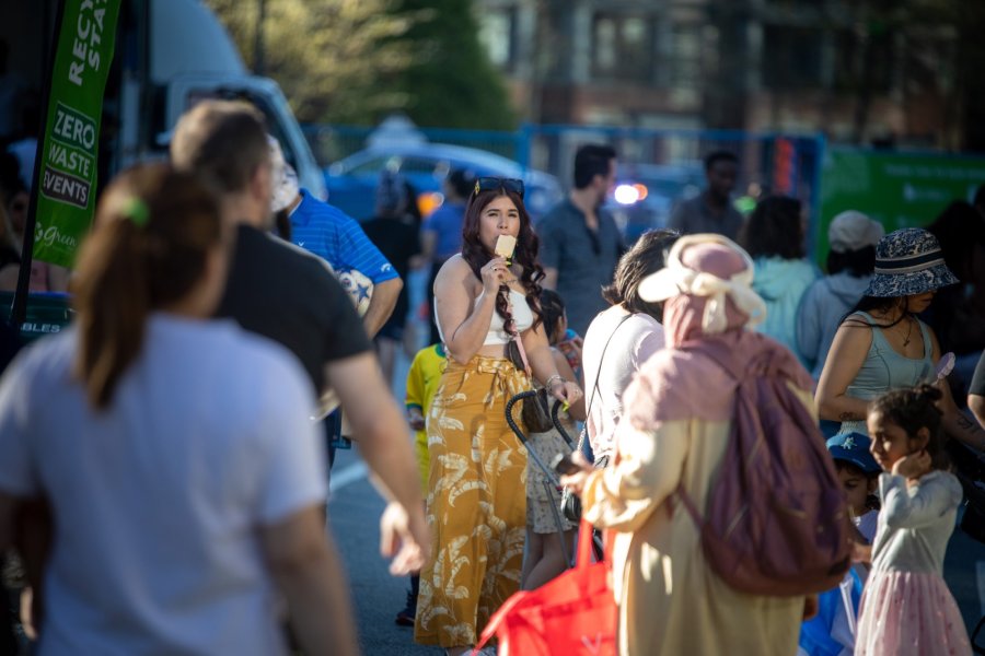 A woman eating ice cream amidst a bustling outdoor event with diverse attendees and a "Zero Waste Events" banner in the background.